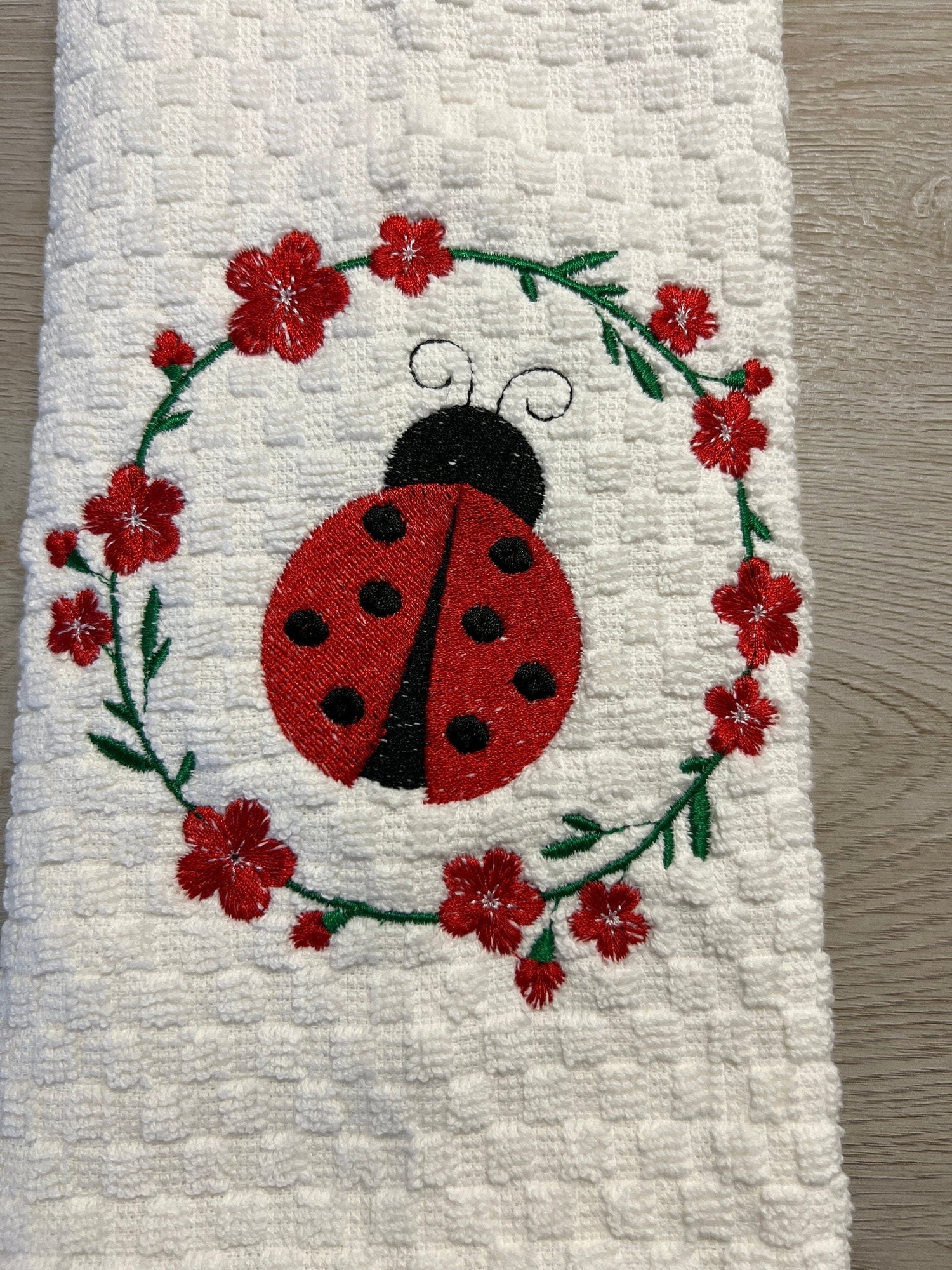 White 100% cotton towel, 15in x 26in, embroidered red ladybug inside a green floral wreath.